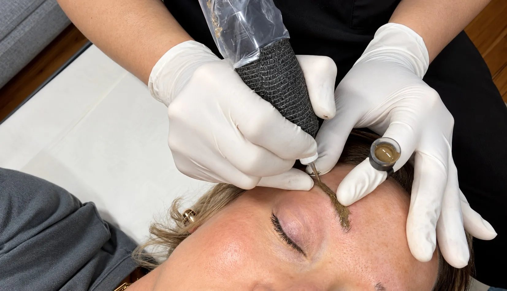 David's gloved hands at work on a blonde client's brow, the machine in one hand and a ring of pigment on the other
