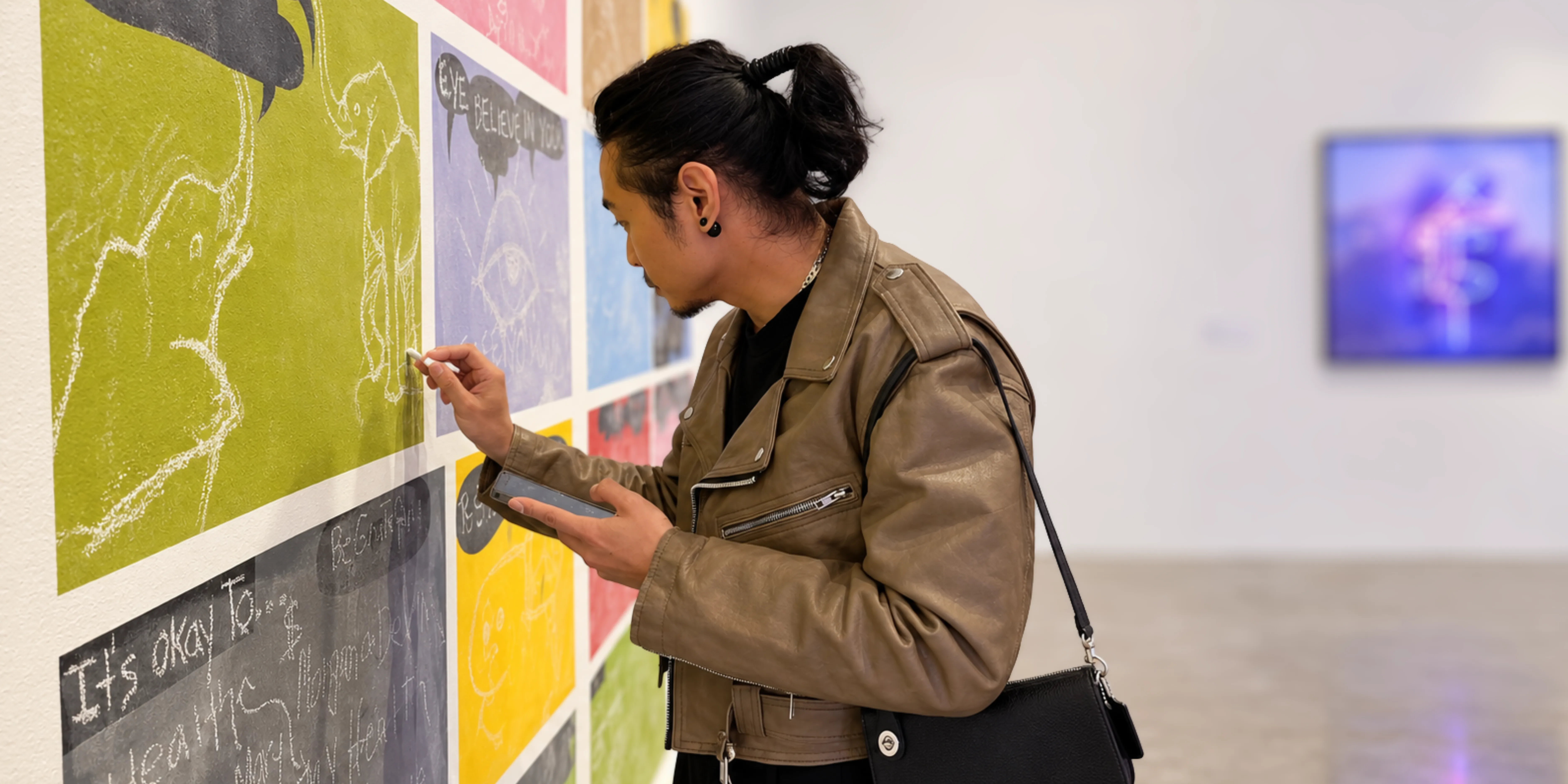 David drawing in chalk on a gallery wall of coloured panels, phone in hand, in a brown leather jacket