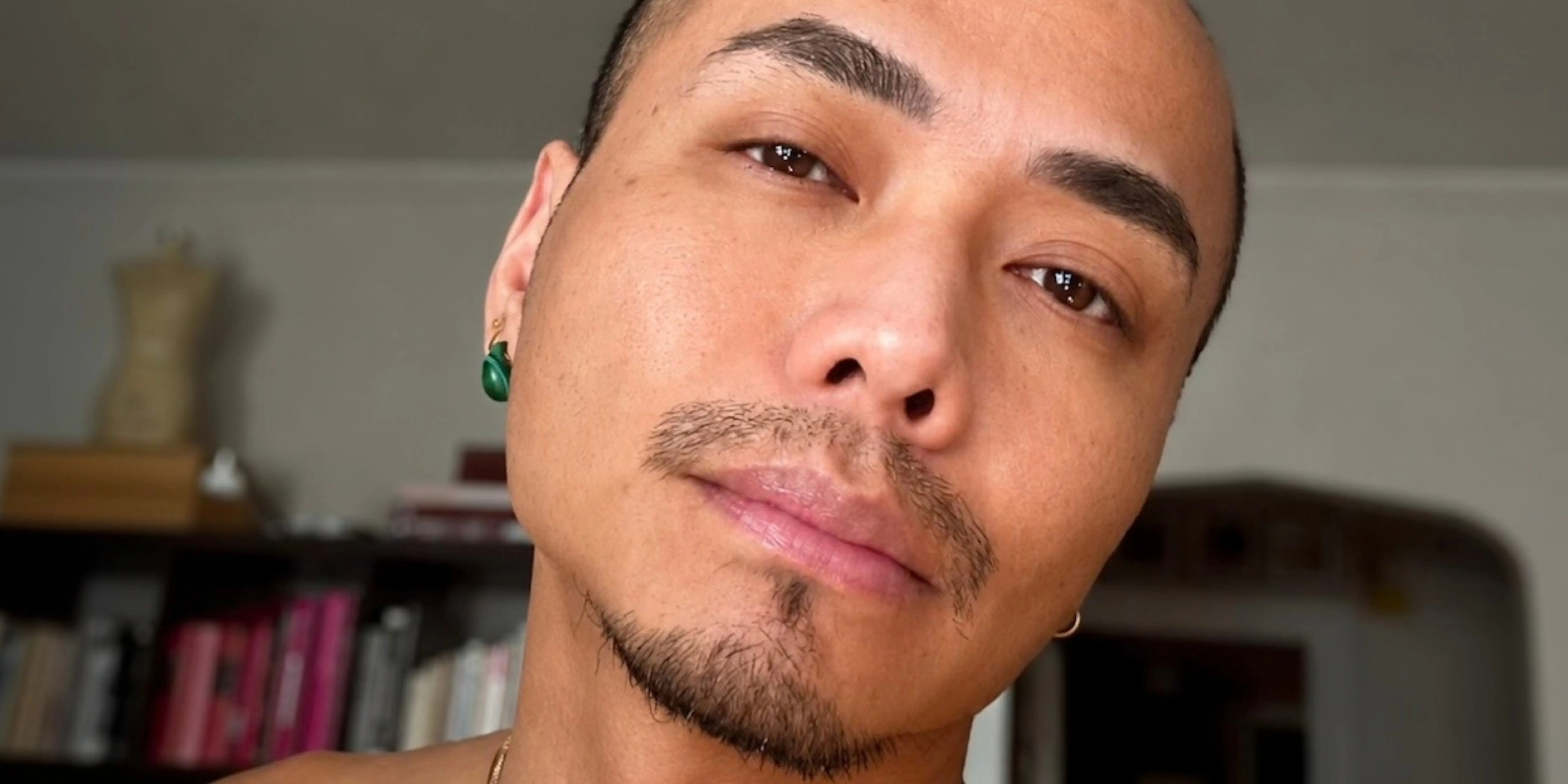 David, a close portrait, his head tilted, a green stone earring, bookshelves behind him