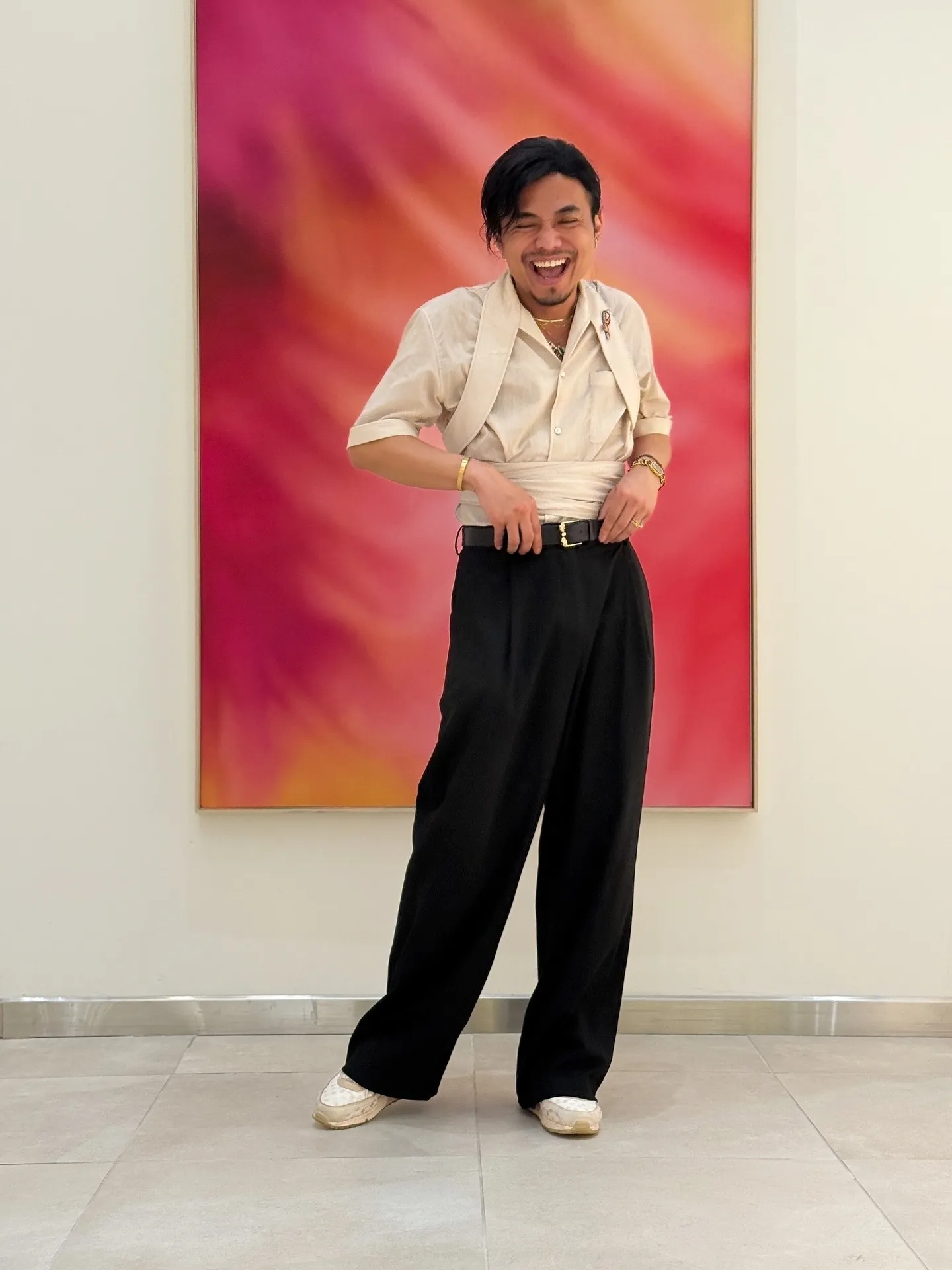 David laughing, in a cream shirt with a sash tied at the waist and wide black trousers, in front of a pink and orange painting