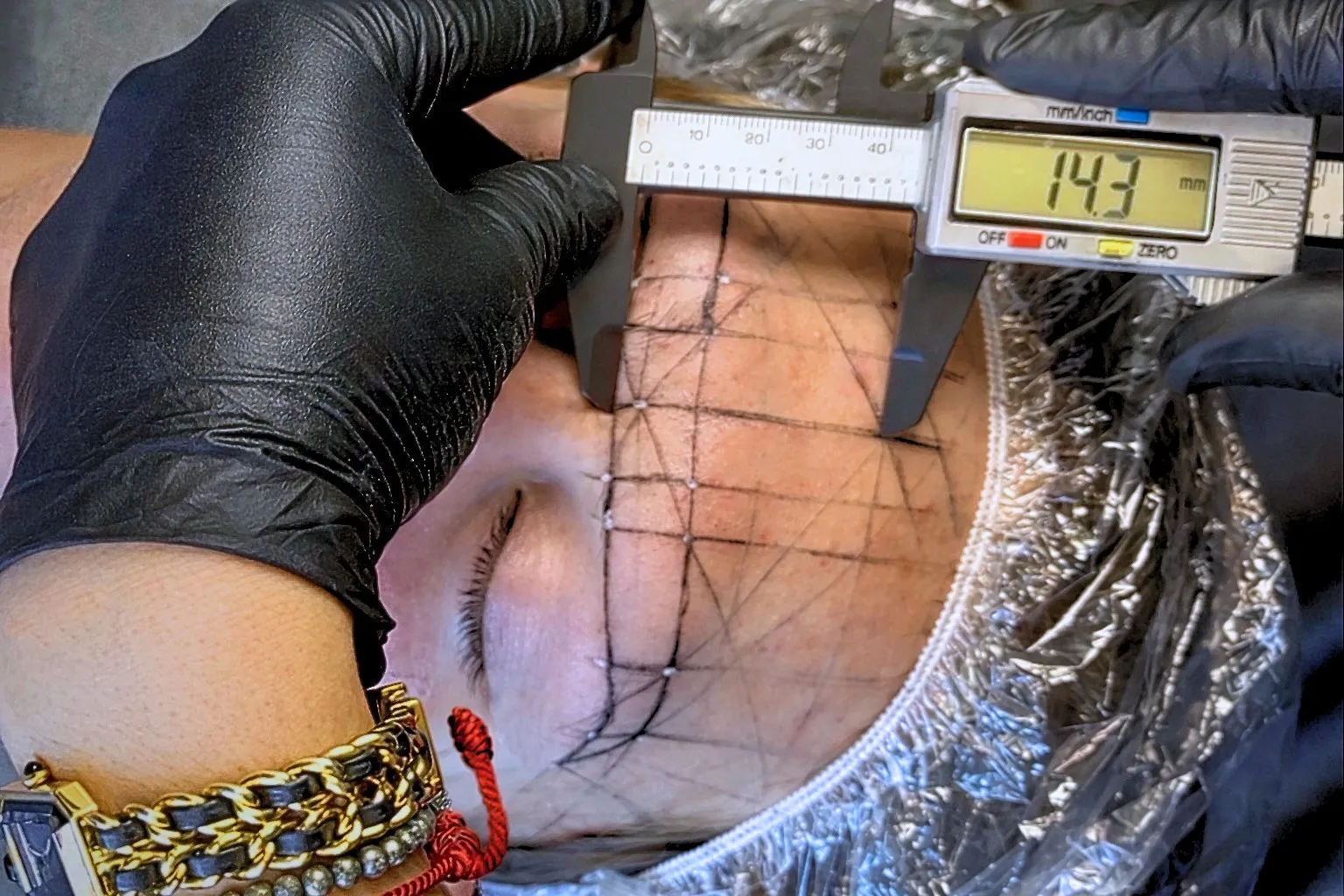 Facial mapping before any tattooing — a caliper measuring the brow across fine mapping lines drawn on a client's forehead