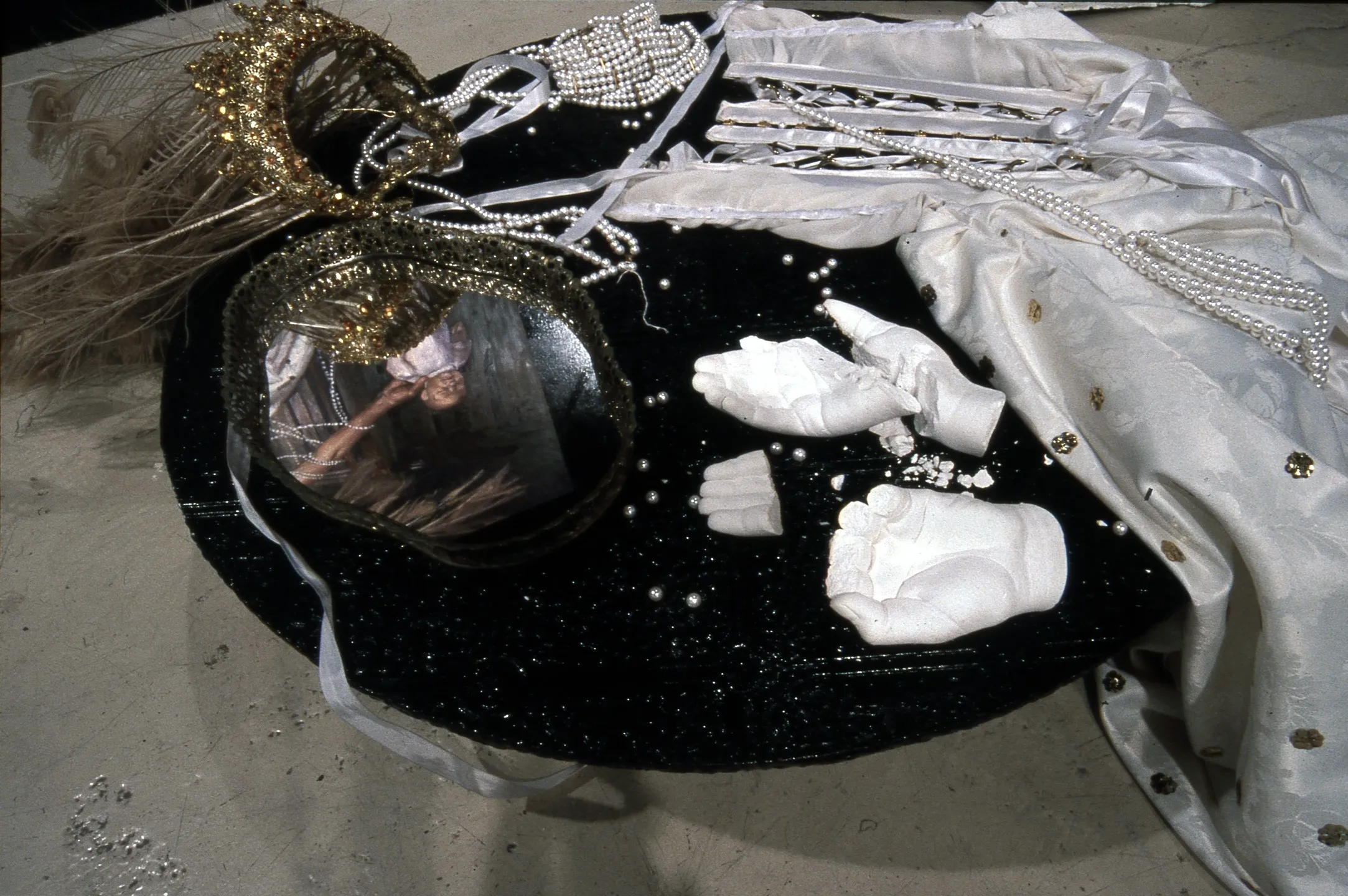 A detail — a gold crown and feathers, strands of pearls, broken plaster hands and a white gown on a black disc