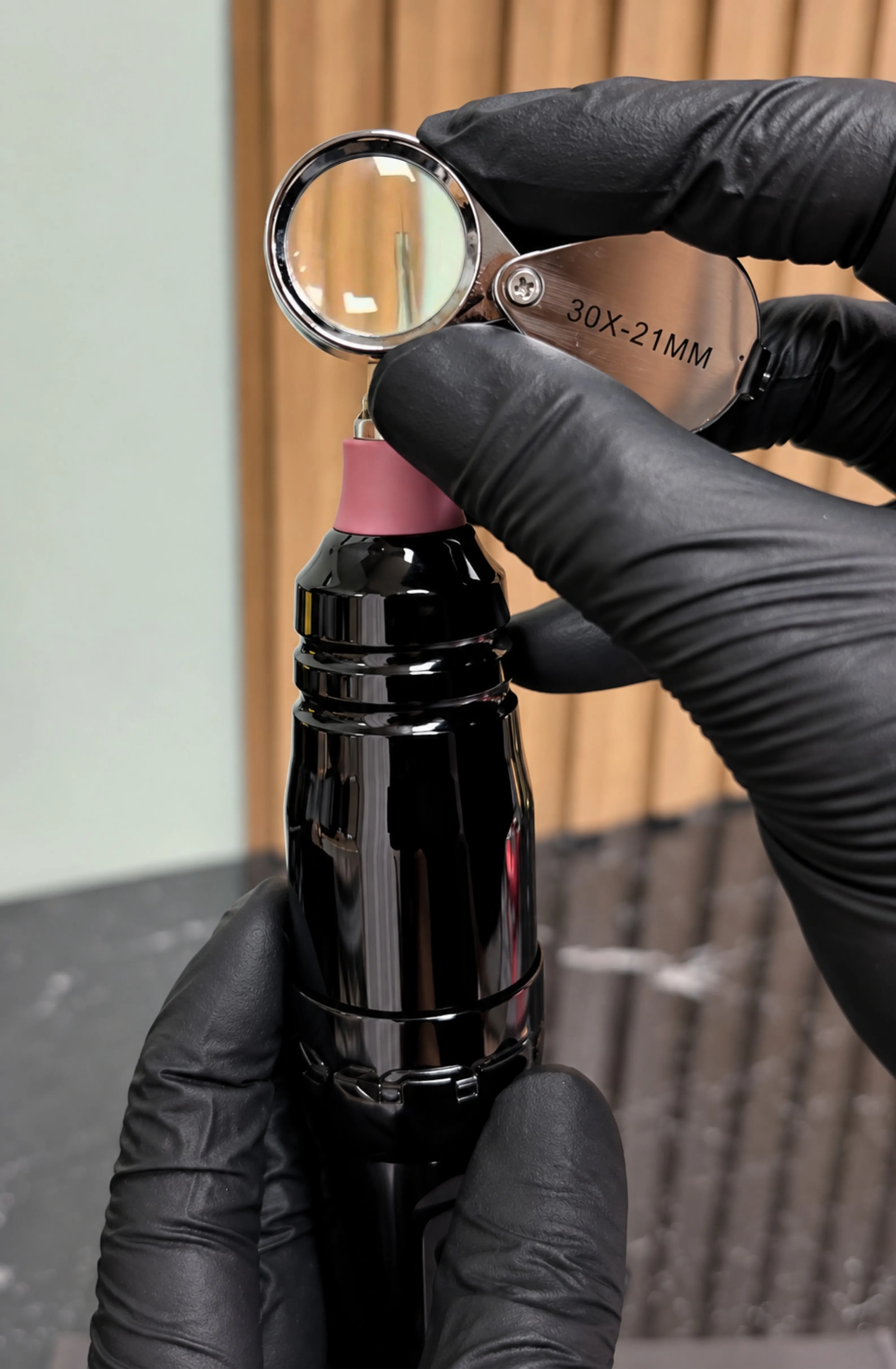 Gloved hands checking a tattoo machine's needle through a 30× loupe
