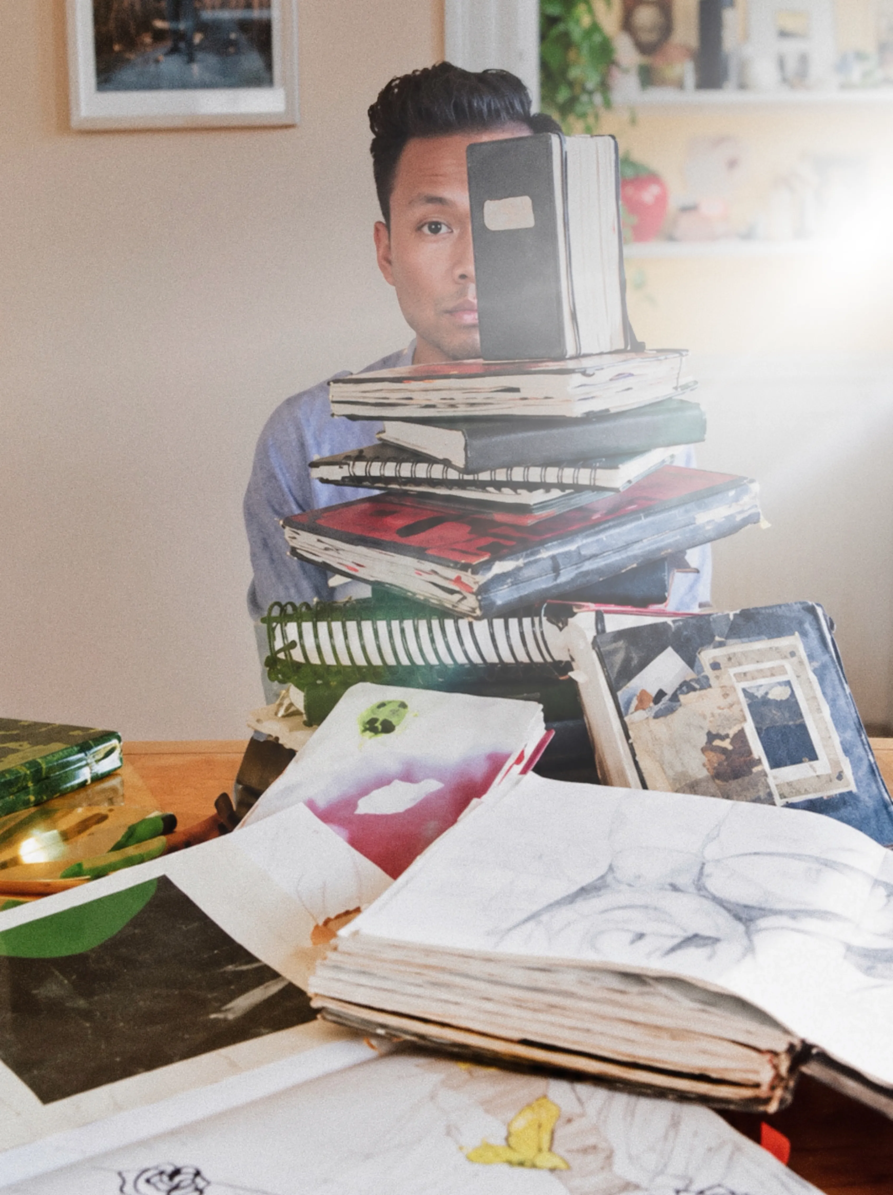 David behind a tall stack of worn notebooks and sketchbooks, one held up over half his face, open sketchbooks spread across the table