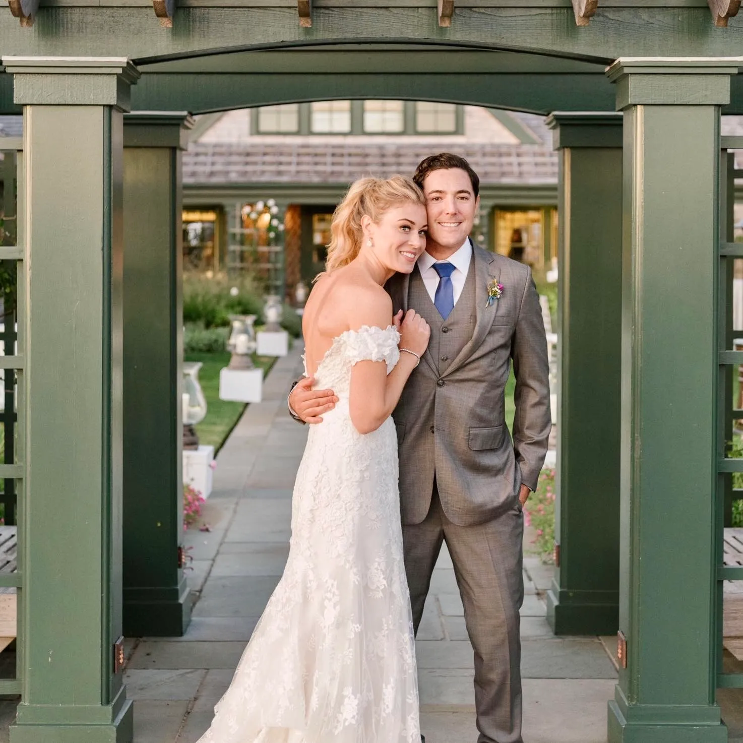 A bride and groom under a green garden pergola — an off-the-shoulder lace gown and a grey three-piece suit with a blue tie