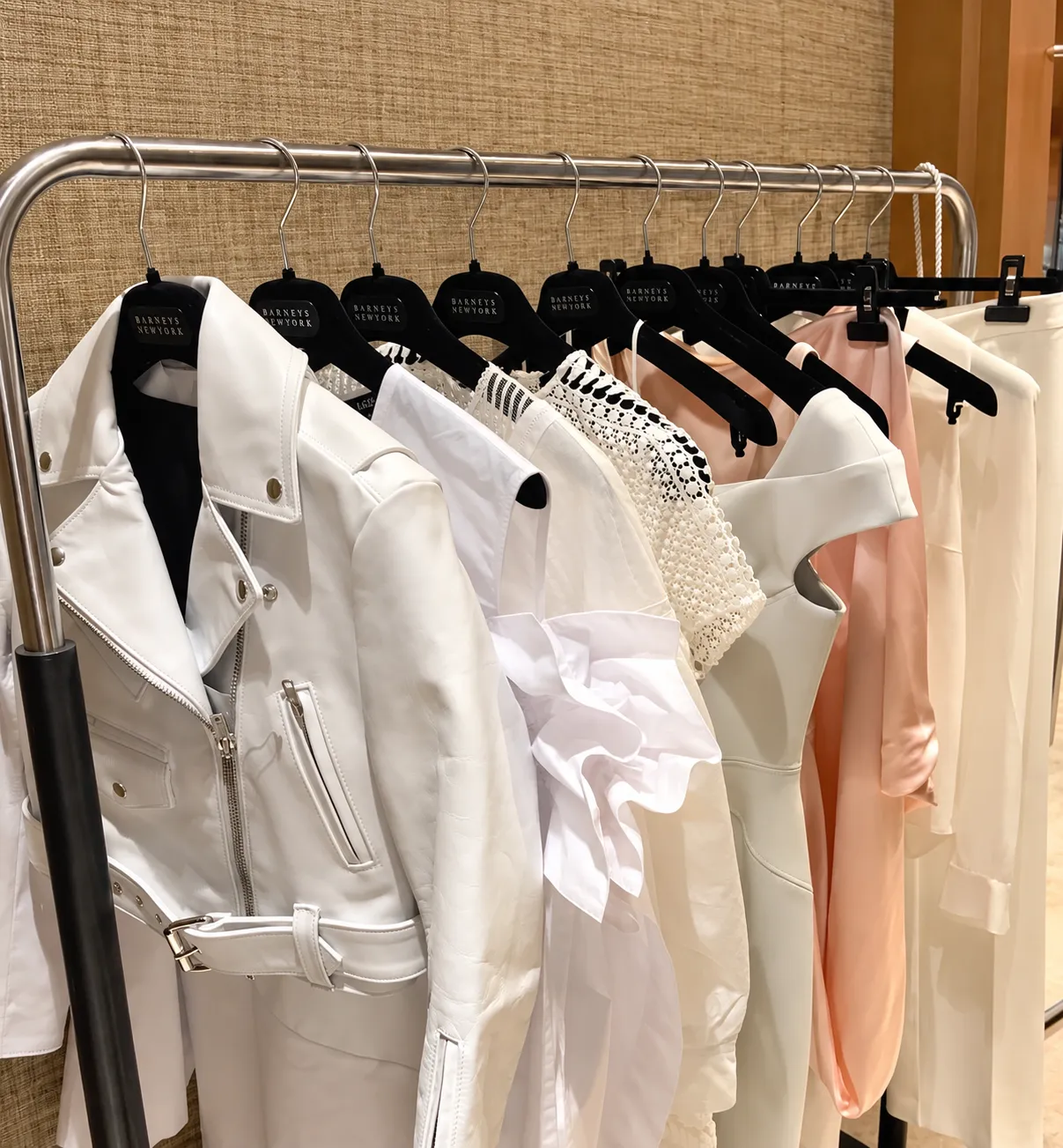 A rail of white and blush pieces on black velvet hangers — a white leather biker jacket, a ruffled white blouse, lace, a white sheath dress and pink silk