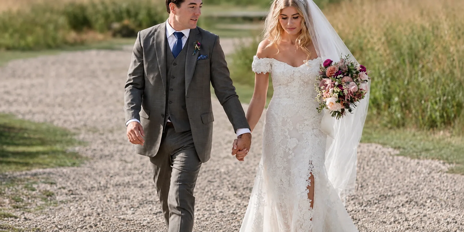 A bride and groom walking hand in hand down a gravel path — a lace off-the-shoulder gown and a grey three-piece suit