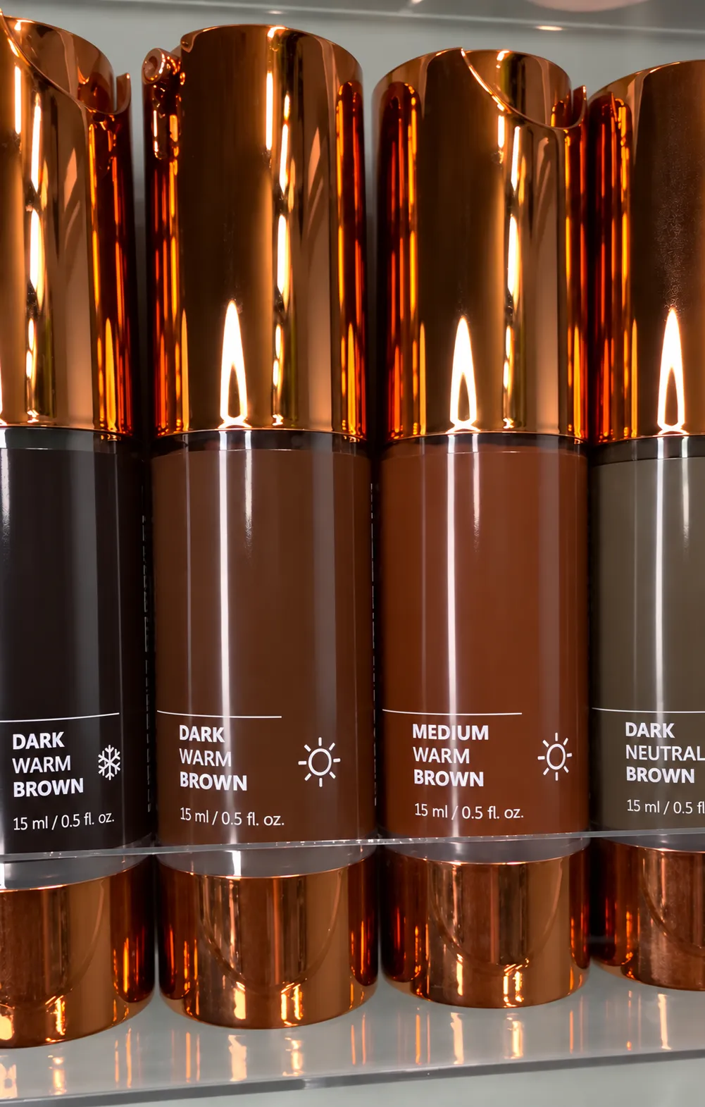 Brow pigments in copper-capped bottles on a glass shelf — Dark Warm Brown, Medium Warm Brown, Dark Neutral Brown