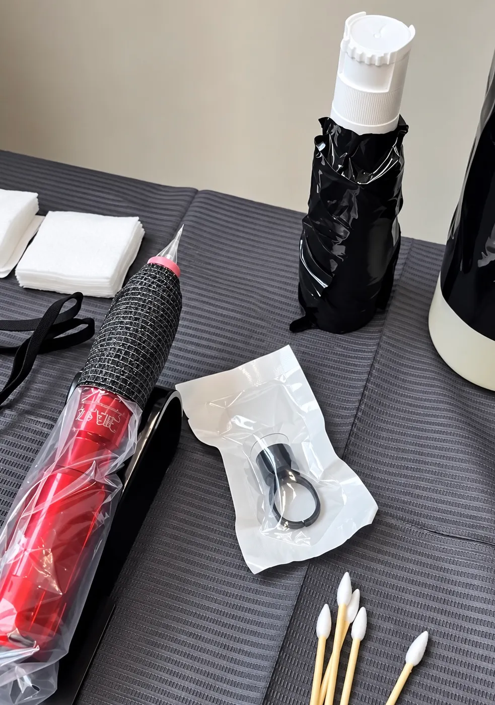 A station set for a touch-up — a wrapped tattoo machine, a sealed pigment ring, gauze and swabs on a grey cover