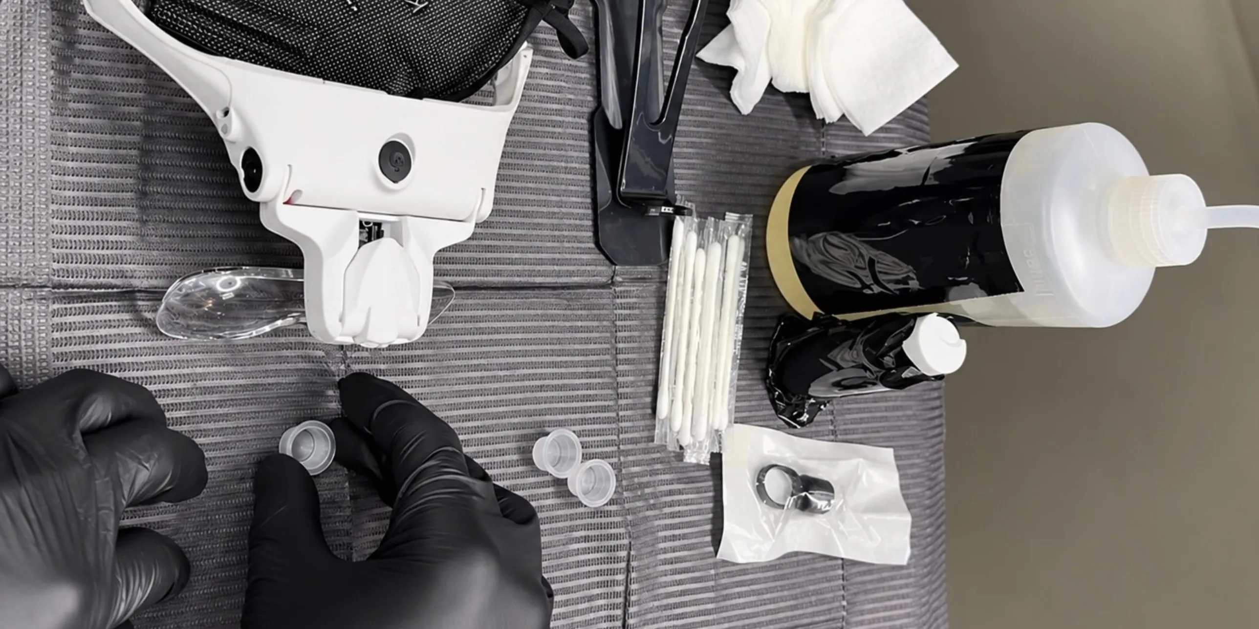 A station laid out for a brow appointment — gloved hands at the pigment cups, a magnifying visor, sealed swabs, a pigment ring and a wash bottle on a grey drape
