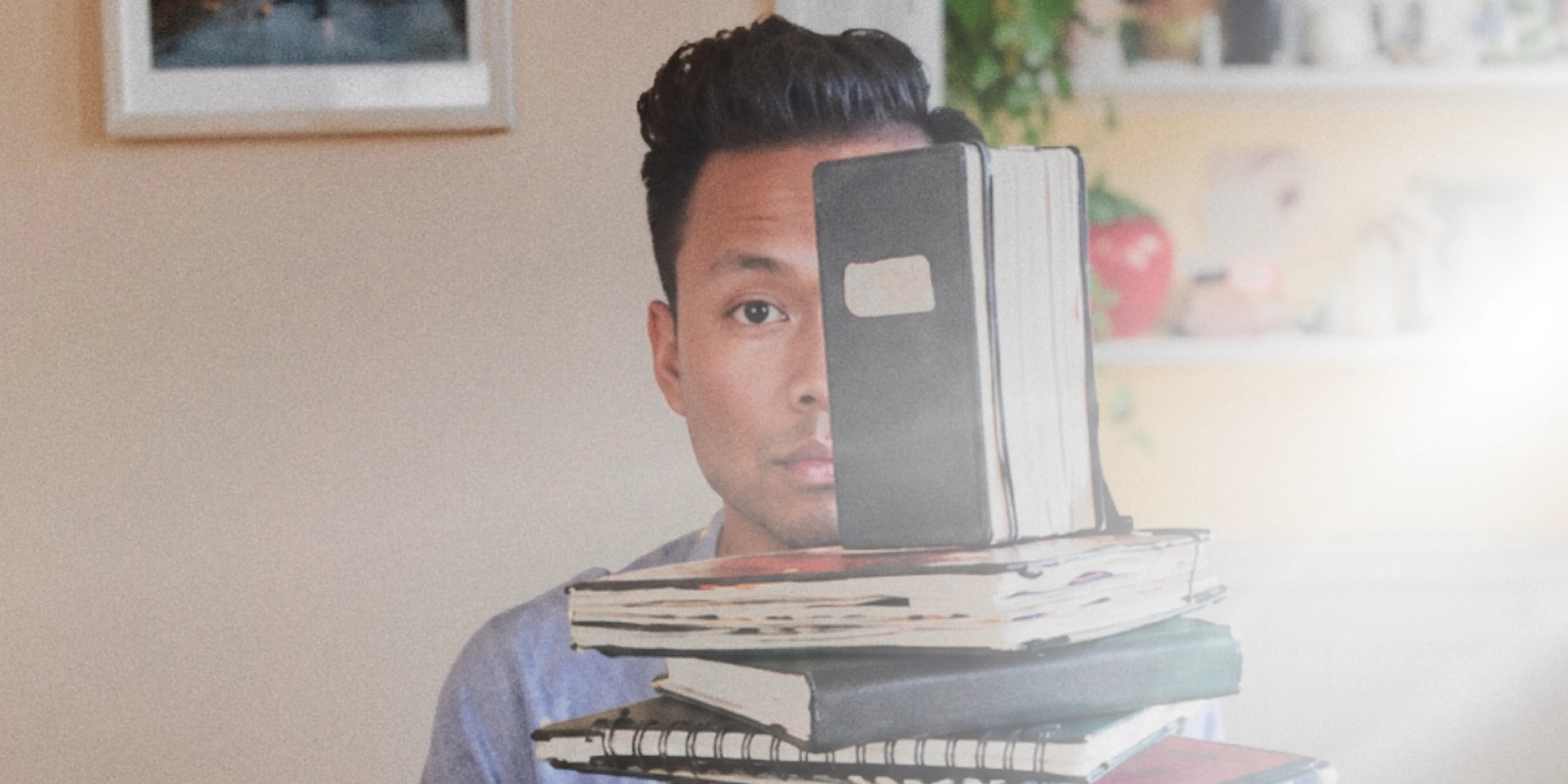 David behind a stack of worn notebooks, one held up over half his face, a framed picture and shelves behind him